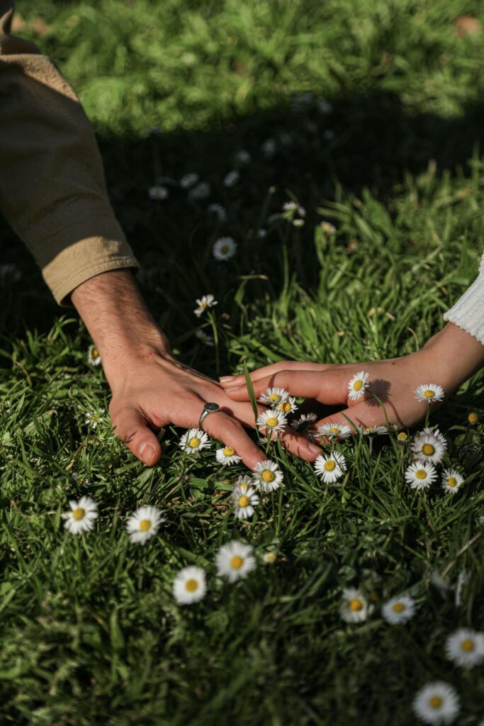 Couple touching hands in grass with white flowers. Begin to understand how communication and intimacy work together to form connections with the help of a relationship therapist in Los Angeles, CA.