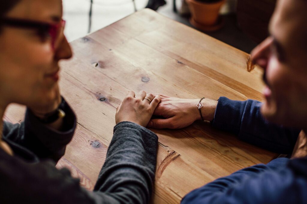 hands | relationship therapy in los angeles, ca | communication in relationships | relationship therapist | Encino | Tarzana | Sherman Oaks Two partners gently holding hands across a wooden table, symbolizing closeness and emotional support. Begin nurturing trust, softness, and emotional safety in your relationship with the help of relationship therapy in Los Angeles, CA.