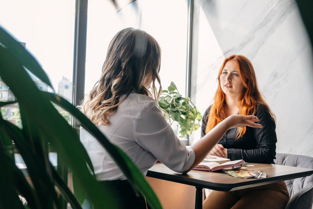 Two women sitting at a table in conversation during a therapy session in a bright office. Build the confidence to communicate authentically with relationship therapy in Los Angeles, CA.