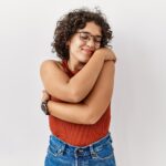 Young woman with curly hair and glasses hugging herself while smiling against a light background. Discover self-compassion and learn to stay emotionally present with yourself through relationship therapy in Los Angeles, CA.