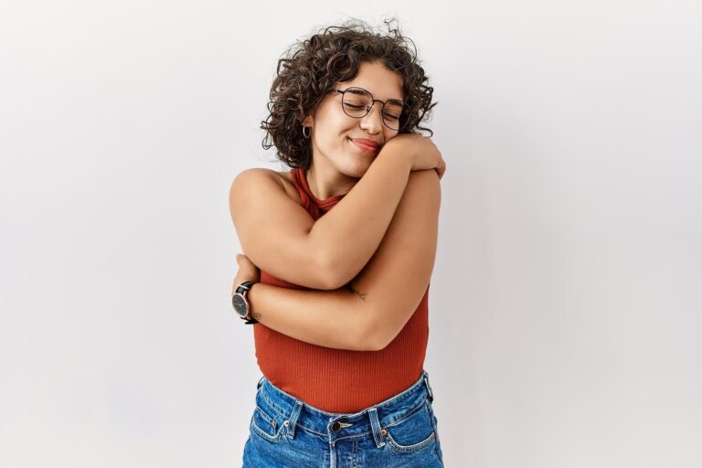 Young woman with curly hair and glasses hugging herself while smiling against a light background. Discover self-compassion and learn to stay emotionally present with yourself through relationship therapy in Los Angeles, CA.