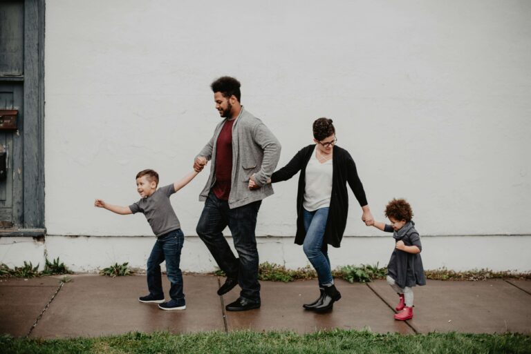 Family of four holding hands while walking together along a sidewalk outdoors. Navigate parenting challenges and stay connected as partners with relationship therapy in Los Angeles, CA.