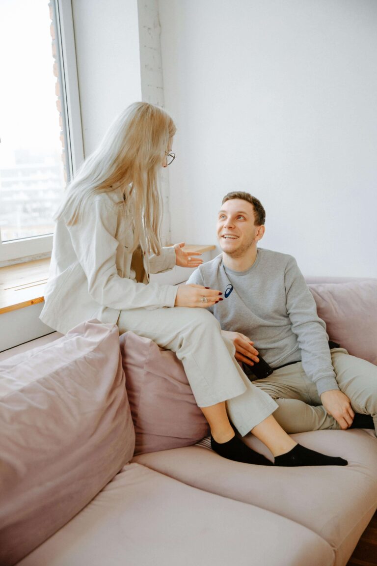 A couple sitting together on a couch, holding hands and smiling at each other in a sunlit room. Learn to trust yourself and deepen your partnership with relationship therapy in Los Angeles, CA.