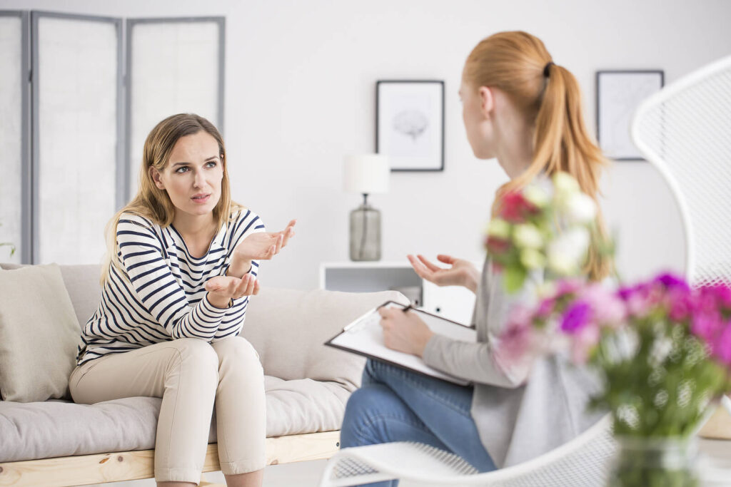Woman in striped shirt sitting on couch gesturing while speaking with therapist taking notes. Understand how your attachment style affects all relationships with relationship therapy in Los Angeles, CA.