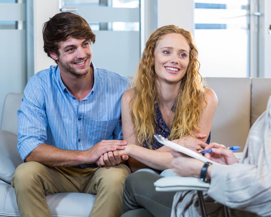 A couple sitting together on a couch smiling while meeting with a therapist. Transform boundary struggles into connection with relationship therapy in Los Angeles, CA.