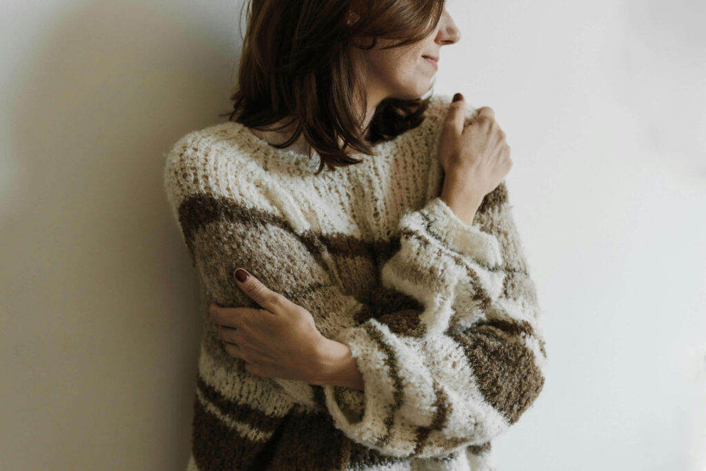 Woman in cozy sweater hugging herself against white wall with gentle smile. Strengthen your relationship with yourself first through relationship therapy in Los Angeles, CA.
