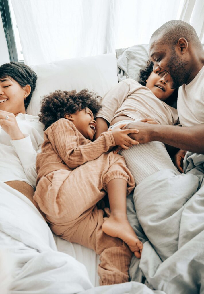 Family of four cuddling and laughing together in bed with natural light. Transform how you connect with your loved ones through relationship therapy in Los Angeles, CA.