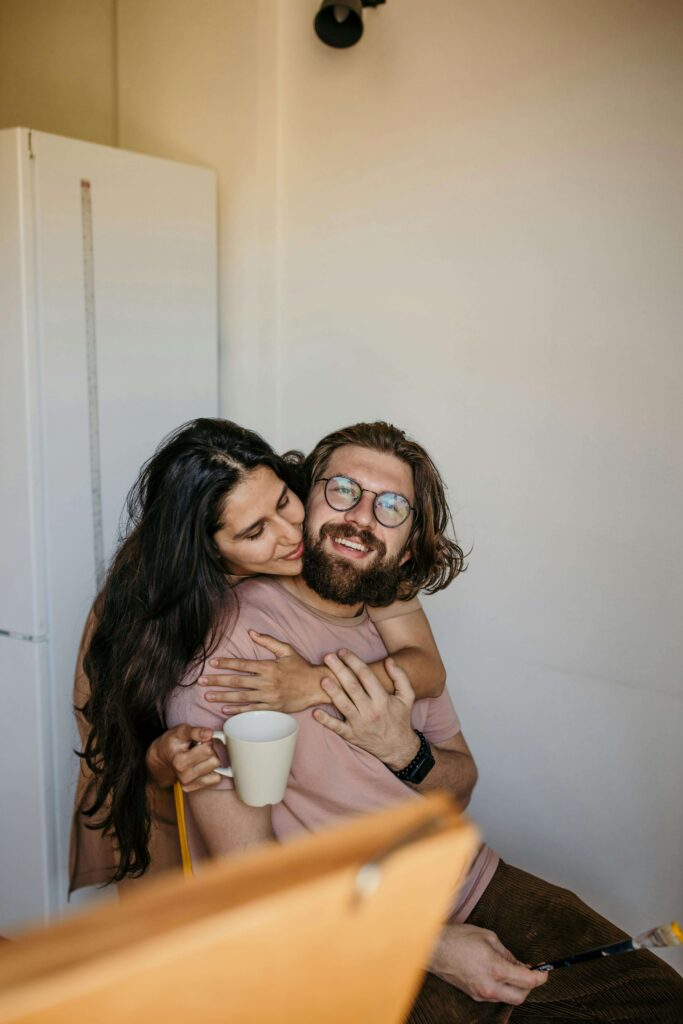 Woman embracing man from behind while he holds coffee mug and smiles. Transform defensiveness into authentic connection through relationship therapy in Los Angeles, CA.