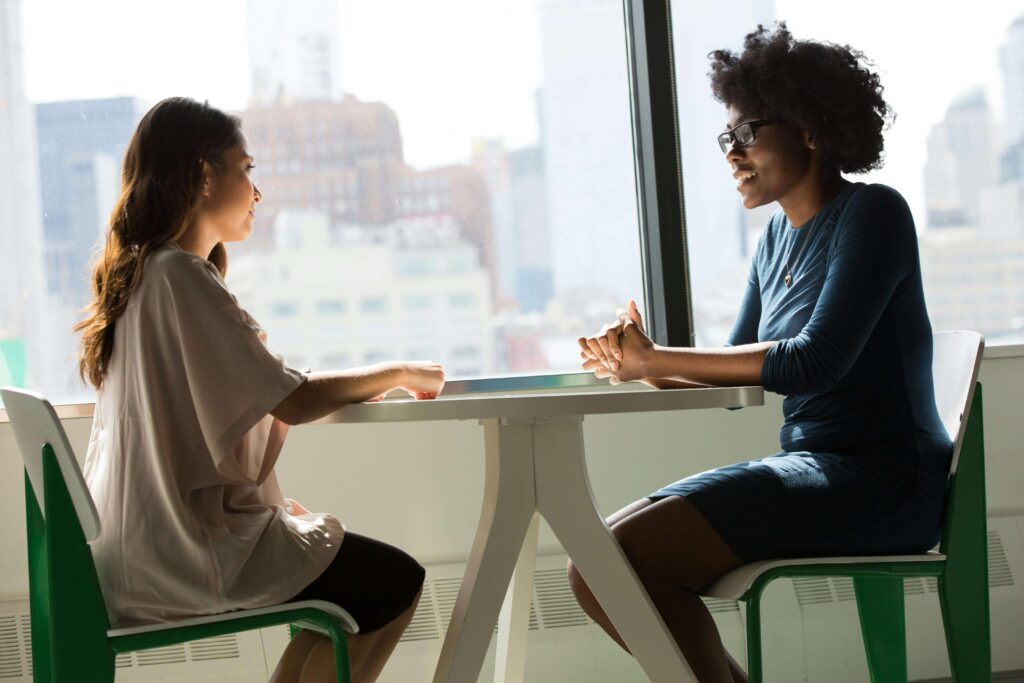 Two women sitting at table facing each other in conversation by window. Explore your inner world to strengthen every relationship through relationship therapy in Los Angeles, CA.