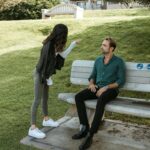 Woman standing and gesturing while man sits on park bench looking up at her. Learn to express pain before it escalates into anger with relationship therapy in Los Angeles, CA.