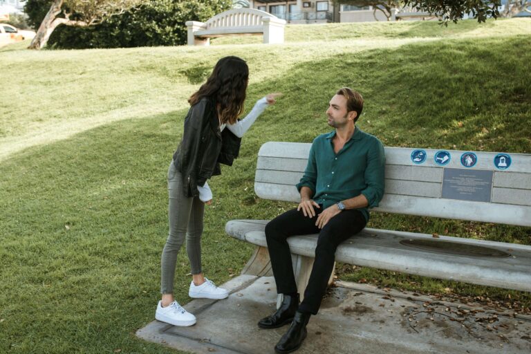 Woman standing and gesturing while man sits on park bench looking up at her. Learn to express pain before it escalates into anger with relationship therapy in Los Angeles, CA.