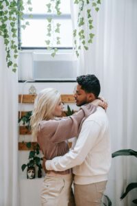 Couple embracing near window with hanging plants in bright home. Move from adapting to being authentic and build real intimacy through relationship therapy in Los Angeles, CA.