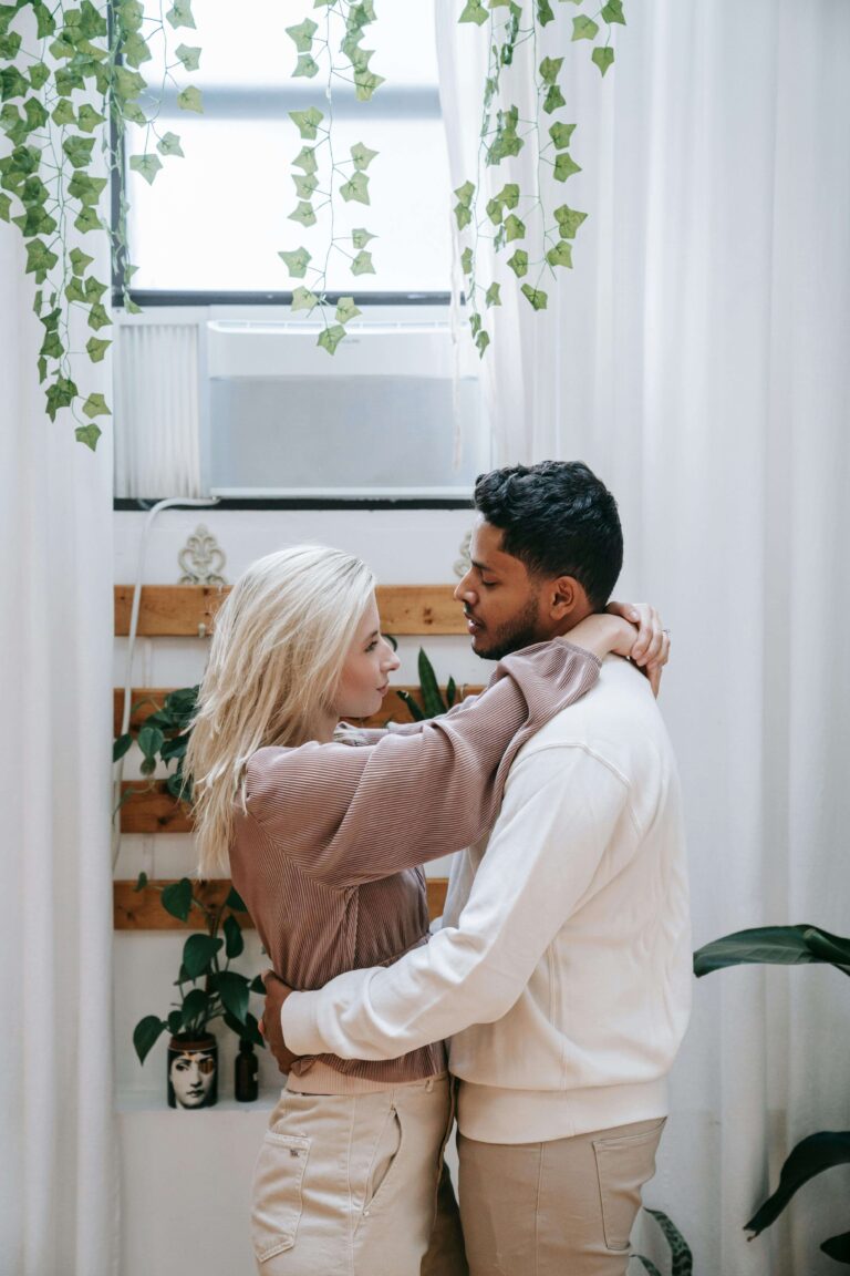 Couple embracing near window with hanging plants in bright home. Move from adapting to being authentic and build real intimacy through relationship therapy in Los Angeles, CA.