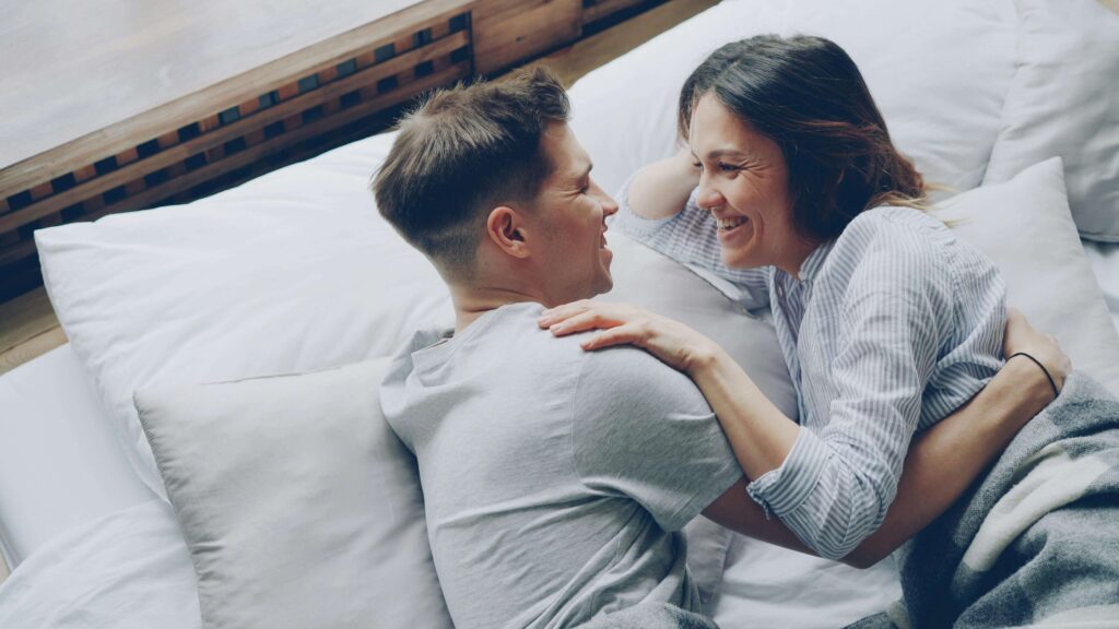happy couple in bed | relationship therapy in Los Angeles, CA | communication in relationships | relationship therapist | West Hills | Agoura Hills | Sherman Oaks Couple lying on bed facing each other smiling and embracing. Rebuild connection with intention and empathy through relationship therapy in Los Angeles, CA.